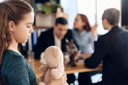 A child, clutching a small toy, sits with their parents in a divorce lawyer's office while the lawyer explains the terms of the child custody agreement.