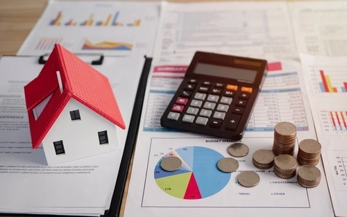 A house model sits on one side of a desk, symbolizing marital property, while on the other side, stacks of cash and a calculator represent the pursuit of a fair division